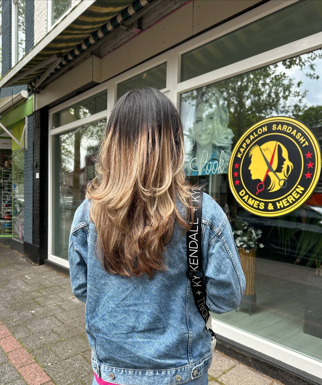 Lang haar met warme blonde lagen, voor het raam van Kapsalon Sardasht met de ronde sticker 'Dames & Heren'